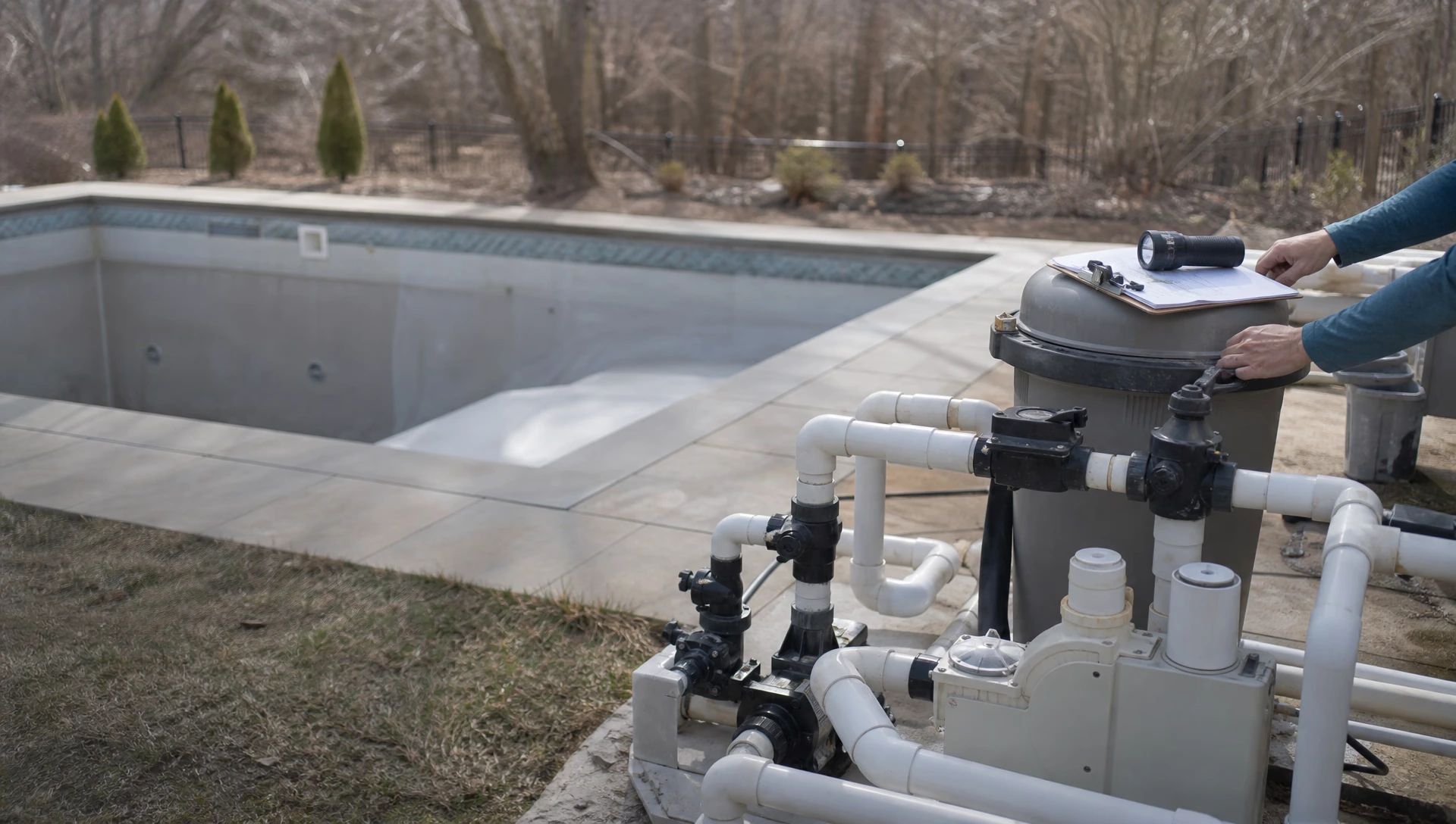 A dry pool equipment pad in early spring with a torch and clipboard resting on the filter, the pool empty of water behind