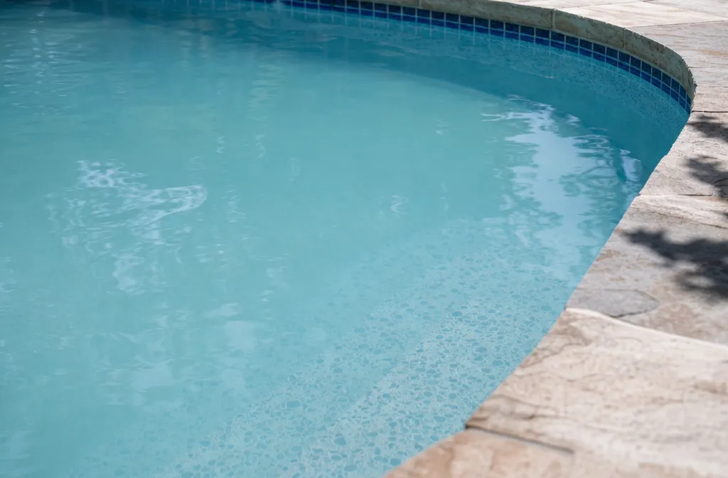 An in-ground pool in bright hot weather where the water has gone faintly milky, the floor pattern soft-edged in the deep end