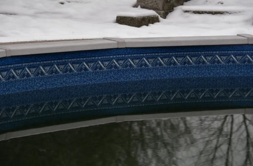 An in-ground vinyl pool in winter with the liner's waterline band visible above dark still water