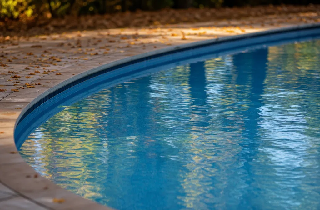 An in-ground pool still open in late September with clear water, low warm light and the first fallen leaves on the deck